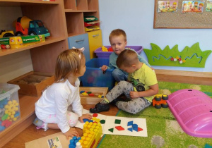 Troje dzieci siedzi na dywanie i układa kompozycje z mozaiki